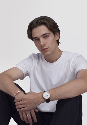 Jonge man met bruin haar, een wit T-shirt, zwarte broek, een polshorloge met een wijzerplaat in wit, en een koperen armband en ring.