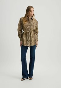 Luipaardprint shirt in beige en bruin, met een riem in de taille, gecombineerd met donkere flared jeans en zwarte sandaaltjes met bandjes. Gladde textuur.
