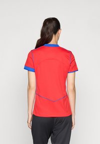 Röd atletisk tröja med korta ärmar, blå krage och detaljer, texturerad tyg och rundade sömmar på baksidan.
