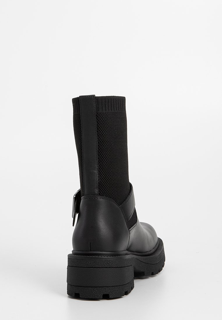 Czarne botki do połowy łydki z masywną podeszwą, dzianinową cholewką, skórzanym obcasem i detalem w postaci paska, widok z tyłu na białym tle.