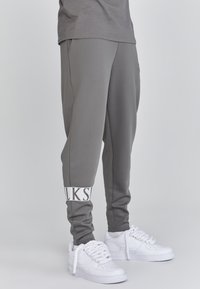 Grijze joggings met zijzakken, taps toelopende pijpen en witte "WALK" branding op de boorden, gecombineerd met witte sneakers met dikke veters.