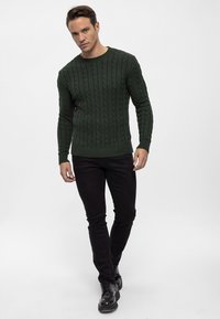 Maglione verde a treccia con collo rotondo, maniche a costine e orlo, abbinato a jeans neri slim-fit e scarpe nere con lacci.
