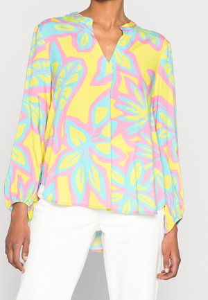 Langærmet bluse med elastiske manchetter, med et farverigt abstrakt blomsterprint i gult, pink og blåt, båret sammen med hvide bukser.