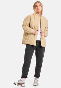 Beige quiltet jakke med snap-front lukking, ribbestrikkede mansjetter og en løs passform, kombinert med svarte bukser og flerfargede joggesko.