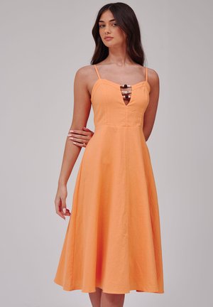 Femme aux longs cheveux foncés portant une robe orange sans manches avec des fines bretelles spaghetti et des perles décoratives au niveau du décolleté.