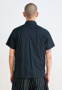 Short-sleeve black shirt with a button-down collar, featuring a smooth texture and a relaxed fit, shown from the back. Striped pants visible.