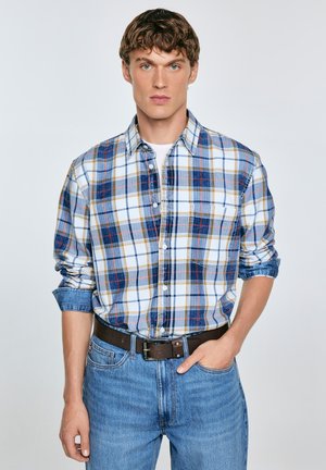 Joven con cabello rizado que lleva una camisa de cuadros azul, blanca y amarilla sobre una camiseta blanca, jeans azules y un cinturón marrón, con una mano en el bolsillo.