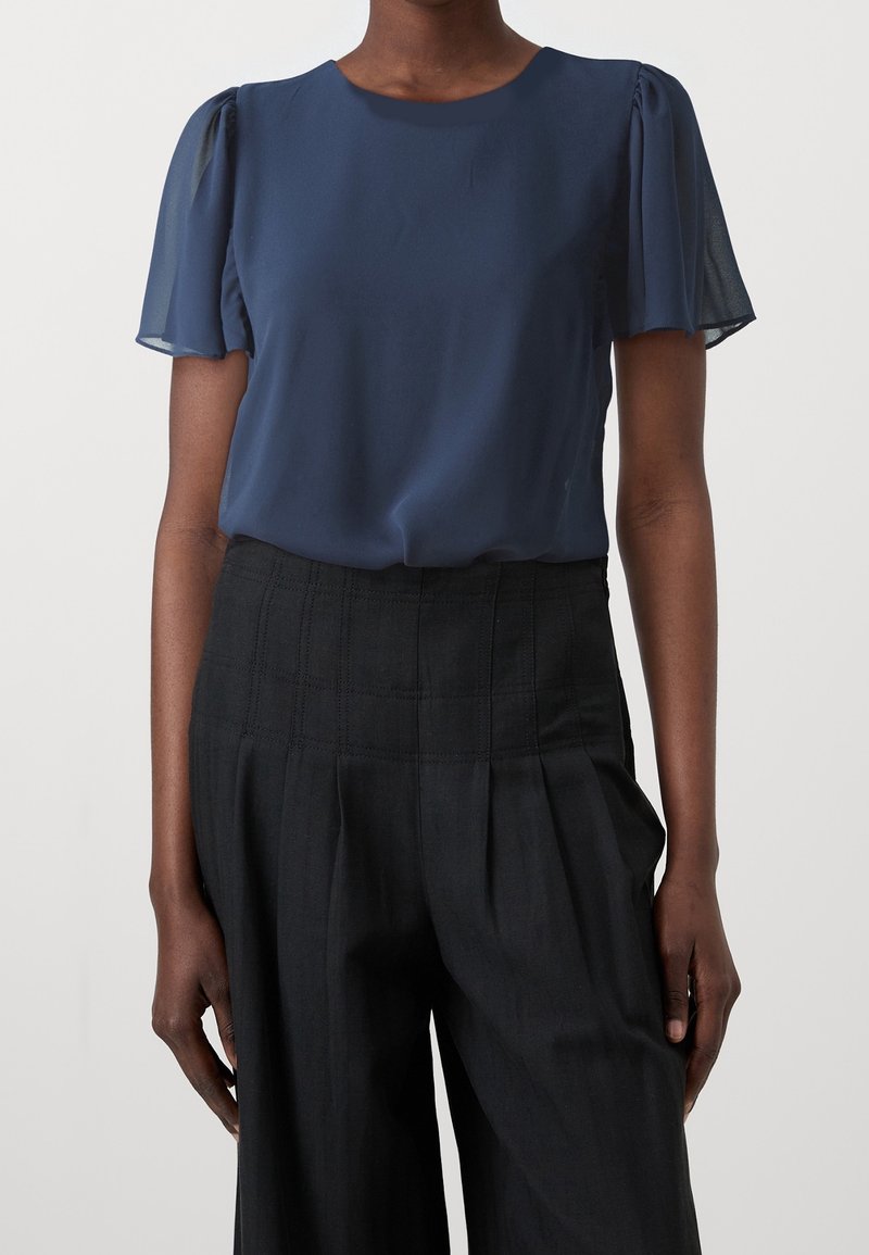 Femme portant un chemisier bleu marine à manches courtes avec des manches bouffantes transparentes et un pantalon noir large taille haute.