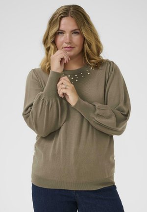 Femme aux cheveux blonds ondulés portant un pull marron avec des détails en perles près de l'encolure, posant les mains près de son visage sur un fond uni.