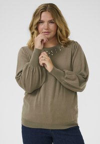 Femme aux cheveux blonds ondulés portant un pull marron avec des détails en perles près de l'encolure, posant les mains près de son visage sur un fond uni.