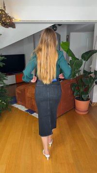 Jupe midi en denim noir avec poches arrière, taille haute et silhouette droite. Portée avec un haut vert et des sandales à talons blanches.