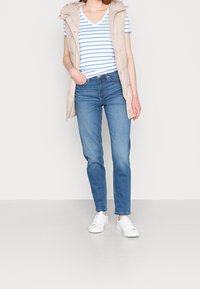 Béžová bezrukávová puffer vesta přes modro-bílé pruhované tričko s výstřihem do V, spárovaná se světle modrými džíny a bílými teniskami.