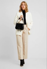 Borsa a tracolla nera con catena, finitura testurizzata, indossata con un blazer bianco, dolcevita nero e pantaloni beige a vita alta, in piedi su scarpe basse.