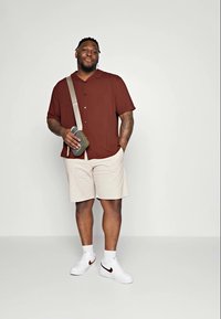 Cămașă maro cu mâneci scurte și nasturi, pantaloni scurți bej, adidași albi cu un swoosh roșu și o geantă crossbody verde. Design simplu și casual.