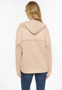 Felpa con cappuccio di colore beige chiaro, dal tessuto morbido, vestibilità ampia e dettagli di cucitura visibili. Presenta una tasca a marsupio e polsini a costine.