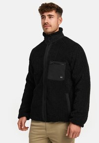 Black fleece jacket with a textured design, zippered pocket on chest, high collar, and smooth zipper detail. Adjusted fit and warm material.