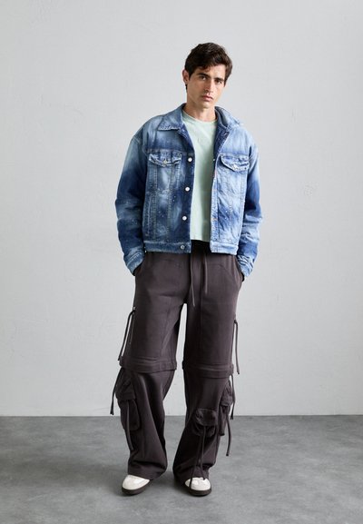 Light blue denim jacket with button-up front, green t-shirt, and dark cargo pants with zippered pockets and drawstring details. White sneakers.