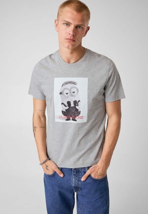 Jeune homme aux cheveux blonds courts portant un t-shirt gris avec un motif Minion et un jean bleu, les mains dans les poches, expression neutre.