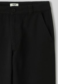 Pantalon noir ample en tissu texturé, doté d'une ceinture boutonnée et de poches latérales, avec un détail de couture minimal.