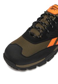 Detailní záběr hnědého a černého teniska Reebok s oranžovým logem a reflexními černými tkaničkami, zobrazující texturované detaily na špičce a podrážce.