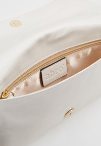 Biała skórzana saszetka o gładkiej fakturze, złote okucia oraz wnętrze wyłożone satyną z kieszenią na zamek i tłoczonym logo.