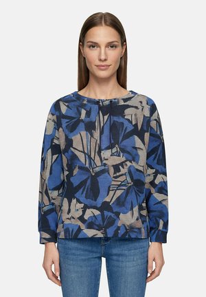 Vrouw met stijl bruin haar die een blauw, zwart en beige bloemetjespatroon lange mouwen top en blauwe spijkerbroek draagt tegen een effen achtergrond.