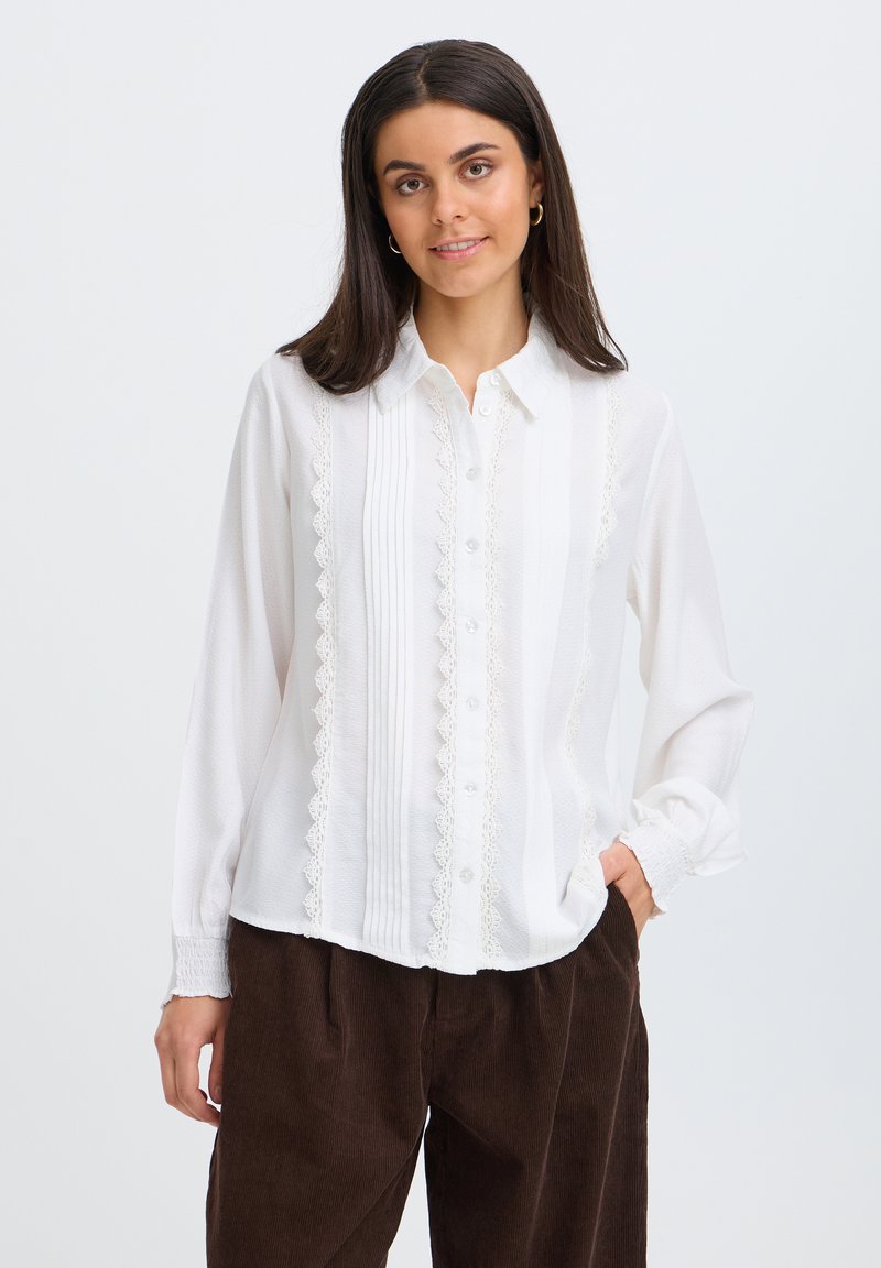 Femme aux cheveux raides et foncés portant un chemisier blanc à manches longues avec des détails en dentelle et un pantalon marron foncé, debout avec une main dans la poche.