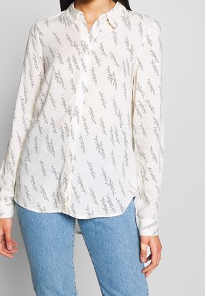 Kvinde iført en hvid langærmet skjorte med knapper og sorte prikmønstre i form af lyn, samt lyseblå jeans, stående med armene afslappede.