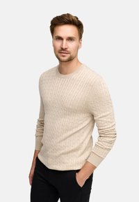 Maglione a trecce beige realizzato in un materiale testurizzato, con scollatura rotonda e polsini a costine, abbinato a pantaloni scuri.