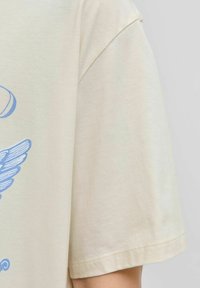 Krátký rukáv, světlé béžové tričko z měkké bavlny, s modrým grafickým designem s motivem křídla na hrudi.