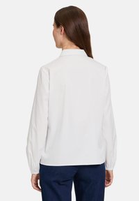 Hvid skjorte med lange ærmer og krave, som har en løs pasform og glat tekstur. Bagsiden er uden udsmykninger og er ensfarvet. Den bæres med mørke bukser.