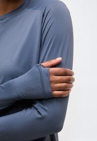 Άτομο που φορά μακρυμάνικο μπλε πουκάμισο με τρύπα για τον αντίχειρα, κρατώντας το αντίθετο άνω χέρι του, σε απλό φωτεινό φόντο.