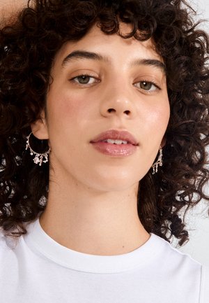 Mladá žena s kučeravými tmavými vlasmi, ktorá má na sebe dekoratívne visiace náušnice kruhy a biely top, pozerá sa mierne nahor s neutrálnym výrazom.
