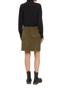 Frau in einem schwarzen Pullover, olivgrüner Cordrock mit Gesäßtaschen und schwarzen Knöchelstiefeln, die von hinten vor einem weißen Hintergrund steht.