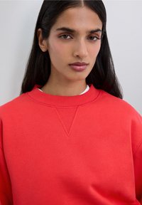 Jeune femme aux longs cheveux noirs portant un sweat-shirt rouge vif, regardant directement l'objectif avec une expression neutre.