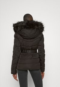 Sort pufferjakke med en halskrave af kunstpels, bælte i taljen og kviltet design med fremtrædende sømme. Glat tekstur, praktisk og varm.