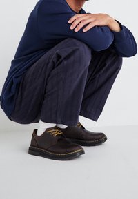 Chaussures en cuir marron avec des lacets et des coutures jaunes, bout rond, semelle en caoutchouc. Portées avec des chaussettes blanches et un pantalon à rayures sombres.