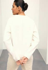 Pull en maille crème avec un col rond, ourlet et poignets côtelés, présentant un motif texturé subtil parcourant le dos. Porté avec un pantalon beige.