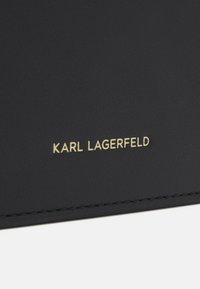 Schwarzes Leder mit strukturiertem Oberflächen, featuring den eingeprägten Namen "KARL LAGERFELD" in goldenen Buchstaben. Gekettelter Rand.