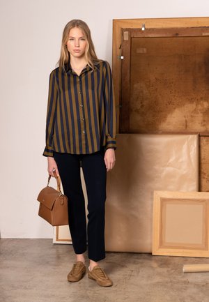 Chemise rayée bleu marine et moutarde, à boutons, associée à un pantalon noir. Sac à main en cuir marron et baskets beiges complètent le look.