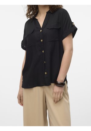 Femme portant une chemise noire ample à manches courtes avec deux poches poitrine et un pantalon beige à jambes larges, debout avec la main droite visible.