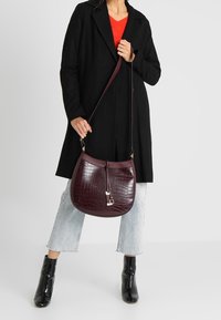 Viininpunainen keinonahkainen olkalaukku krokotiilikuviolla, hopeisilla metalliosilla ja kaarevalla muotoilulla, kantaa olkapäällä ristiin paksun takin ja farkkujen kanssa.