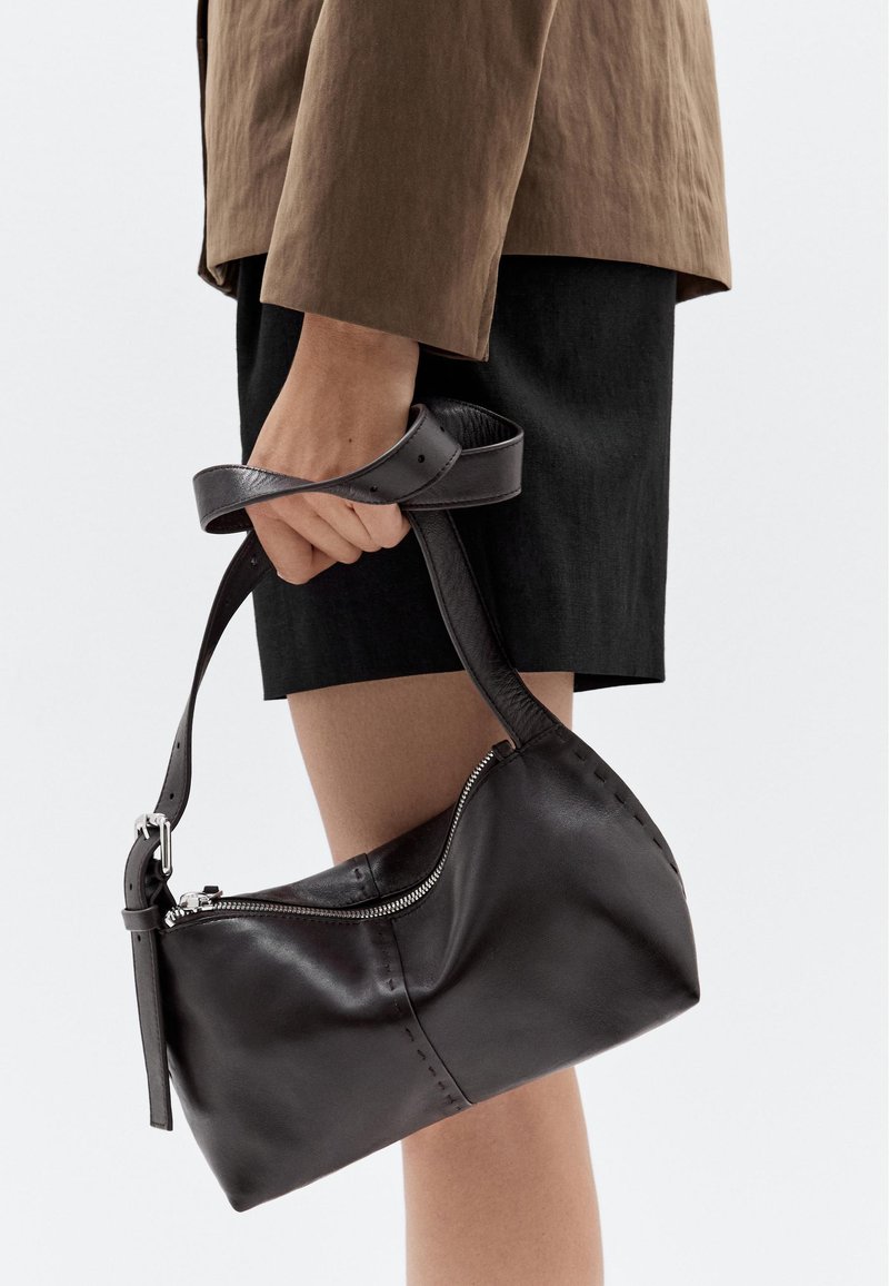 Borsa a tracolla in pelle nera con design curvo, chiusura con zip e tracolla regolabile. La cucitura visibile sui lati aggiunge texture.