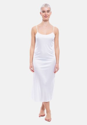 Junge Person mit kurzen Haaren, die ein weißes ärmelloses Satinkleid trägt und barfuß vor einem schlichten hellen Hintergrund steht.