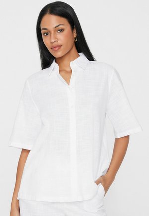 Frau mit langen schwarzen Haaren trägt ein lockeres weißes Hemd mit kurzen Ärmeln und steht mit einer Hand in der Tasche vor einem einfarbigen Hintergrund.