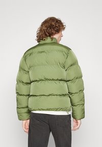 Giubbotto imbottito in verde oliva, con sezioni trapuntate, colletto alto e una texture liscia e lucida. Indossato sopra jeans scuri.