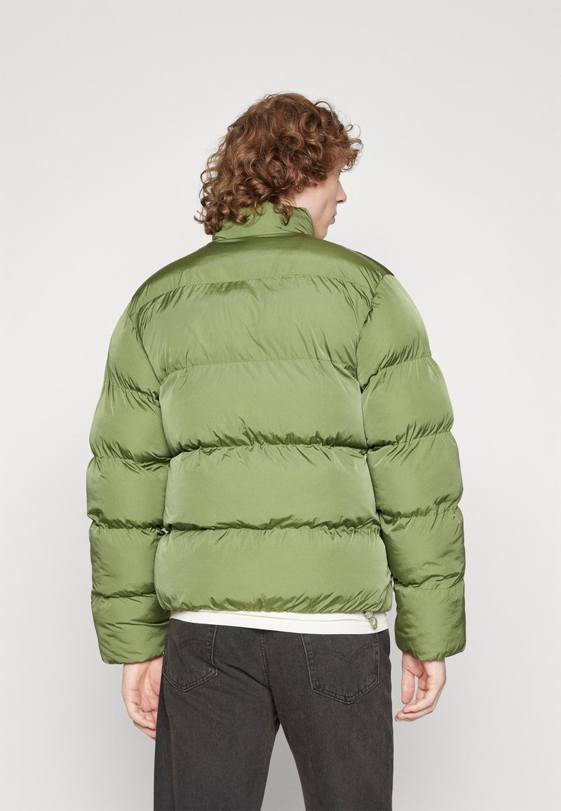 Giubbotto imbottito in verde oliva, con sezioni trapuntate, colletto alto e una texture liscia e lucida. Indossato sopra jeans scuri.