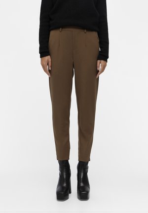 Osoba ubrana w brązowe dopasowane spodnie, czarną bluzkę z długim rękawem oraz czarne botki na wysokim obcasie z platformą, stojąca na tle białego tła.