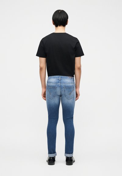 Άνδρας που στέκεται με την πλάτη προς τα εμπρός, φοράει μαύρο μπλουζάκι, ξεβαμμένα στενά τζιν με γυρισμένα μπατζάκια και μαύρα παπούτσια, μπροστά σε απλό λευκό φόντο.