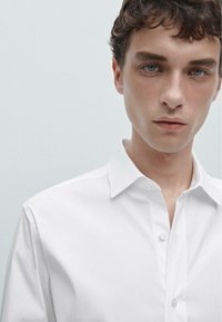 Camicia bianca con colletto a punta, texture liscia e vestibilità classica. Presenta cuciture sottili e chiusura con bottoni standard.